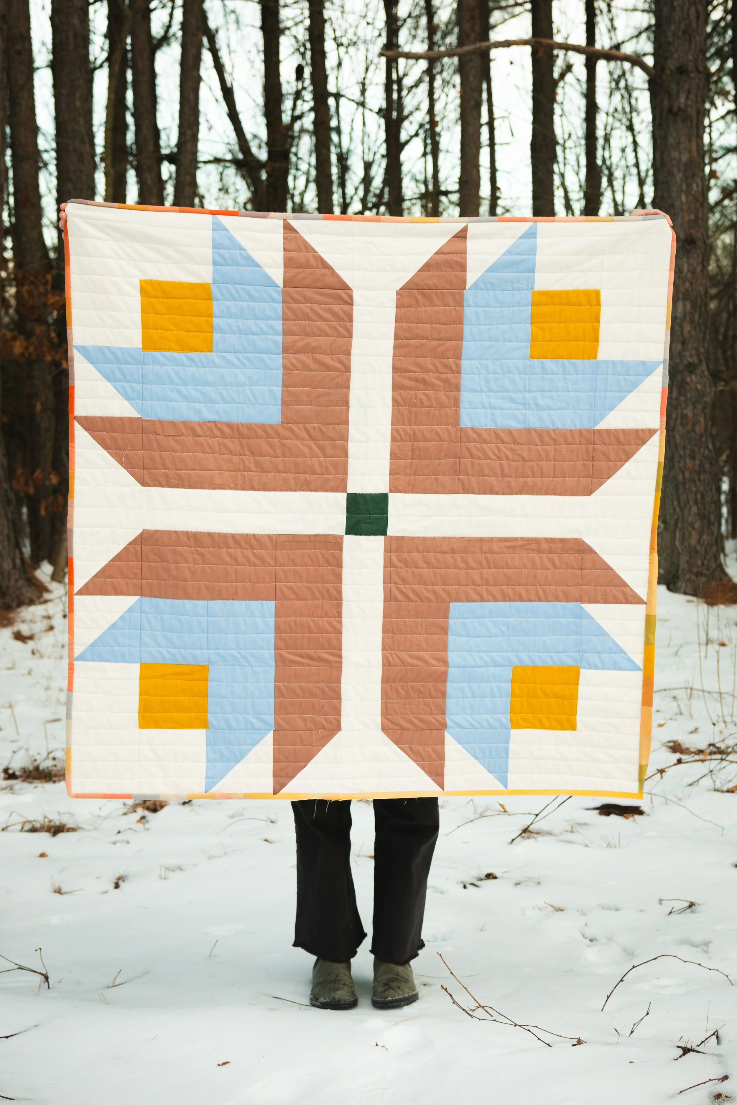 Person holding a quilt with a geometric star pattern in a snowy outdoor setting with trees in the background.