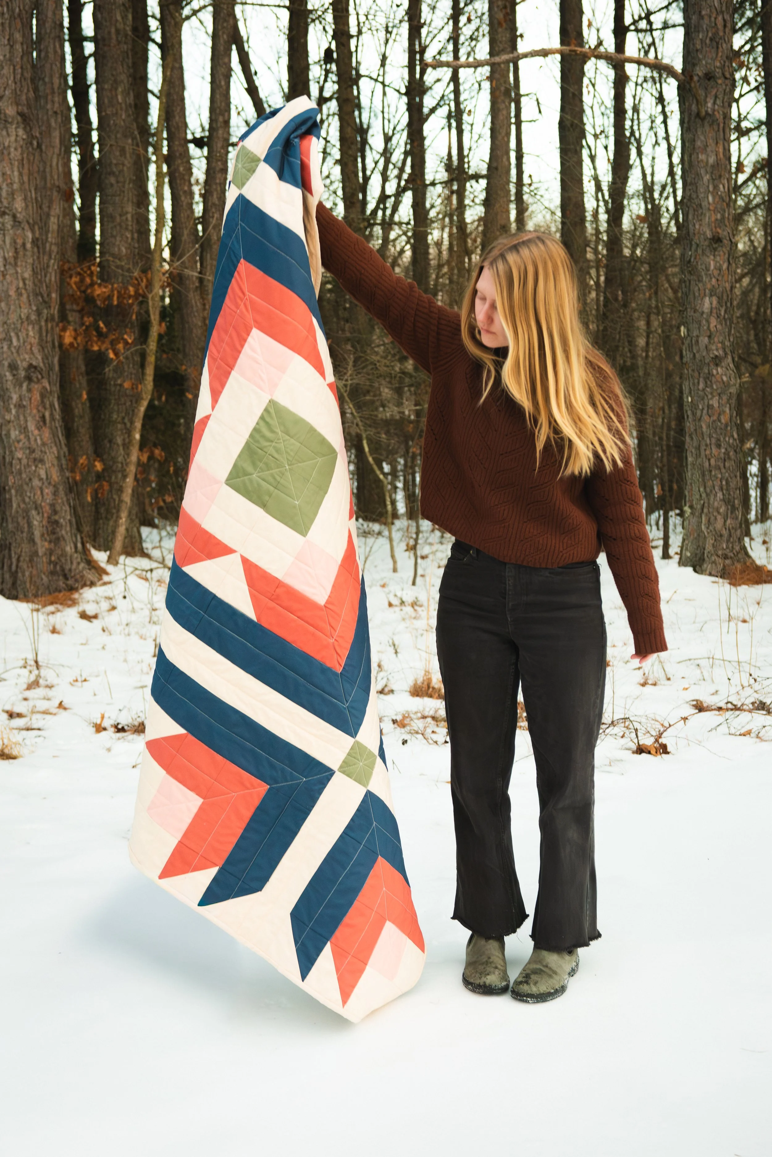 A woman with long blonde hair wearing a brown sweater, black pants, and gray boots, standing outdoors in a snowy forest, holding a colorful quilted blanket with geometric patterns.
