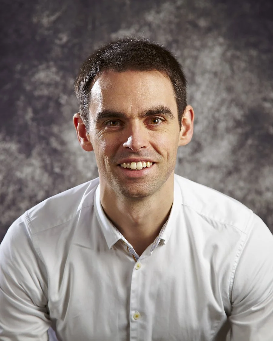 A smiling man with short dark hair wearing a white button-up shirt, sitting against a textured gray background.