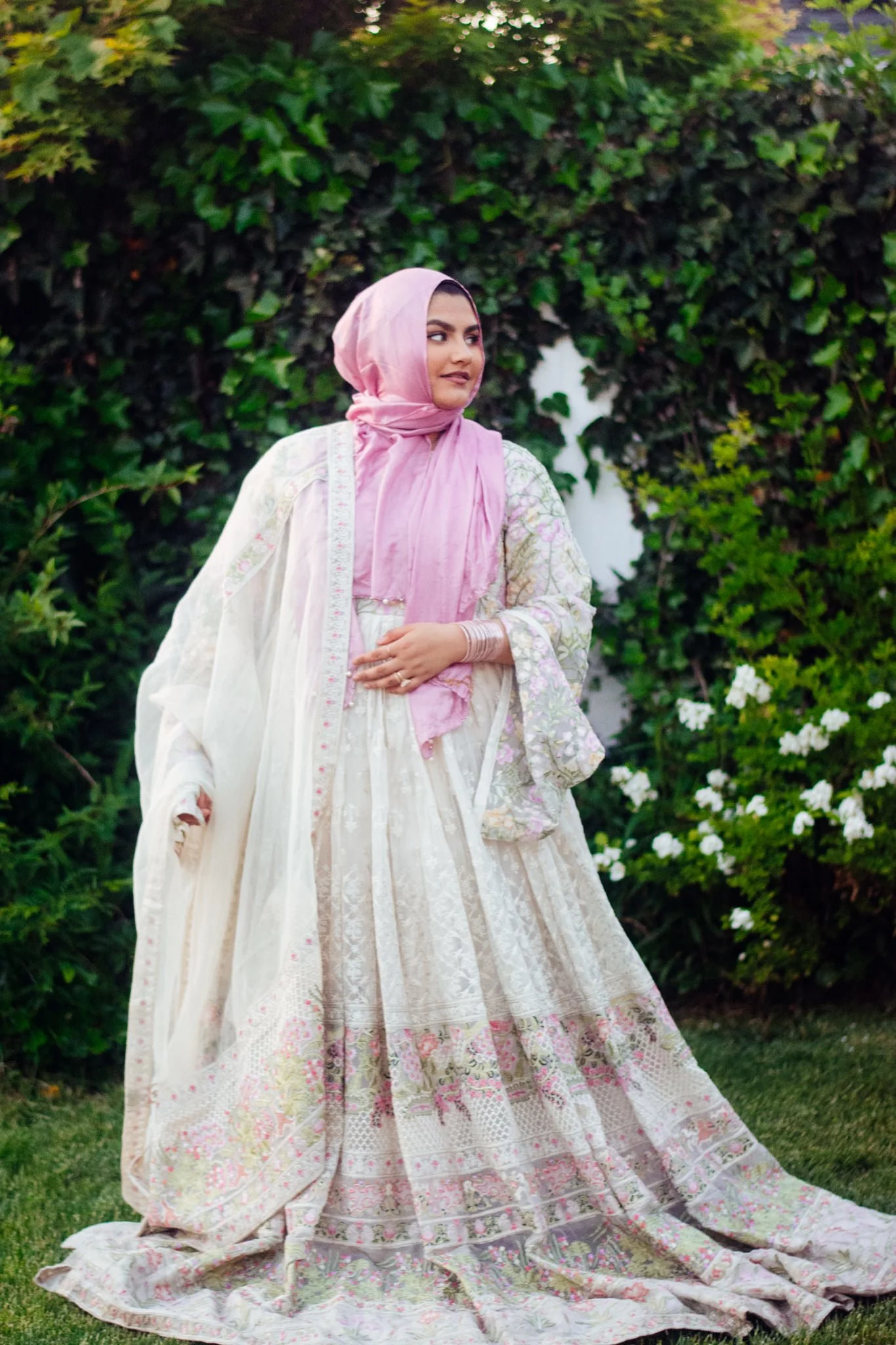 A woman in a traditional, ornate white dress with pink and floral embroidery, wearing a pink headscarf, standing outdoors in front of a leafy green hedge with white flowers.