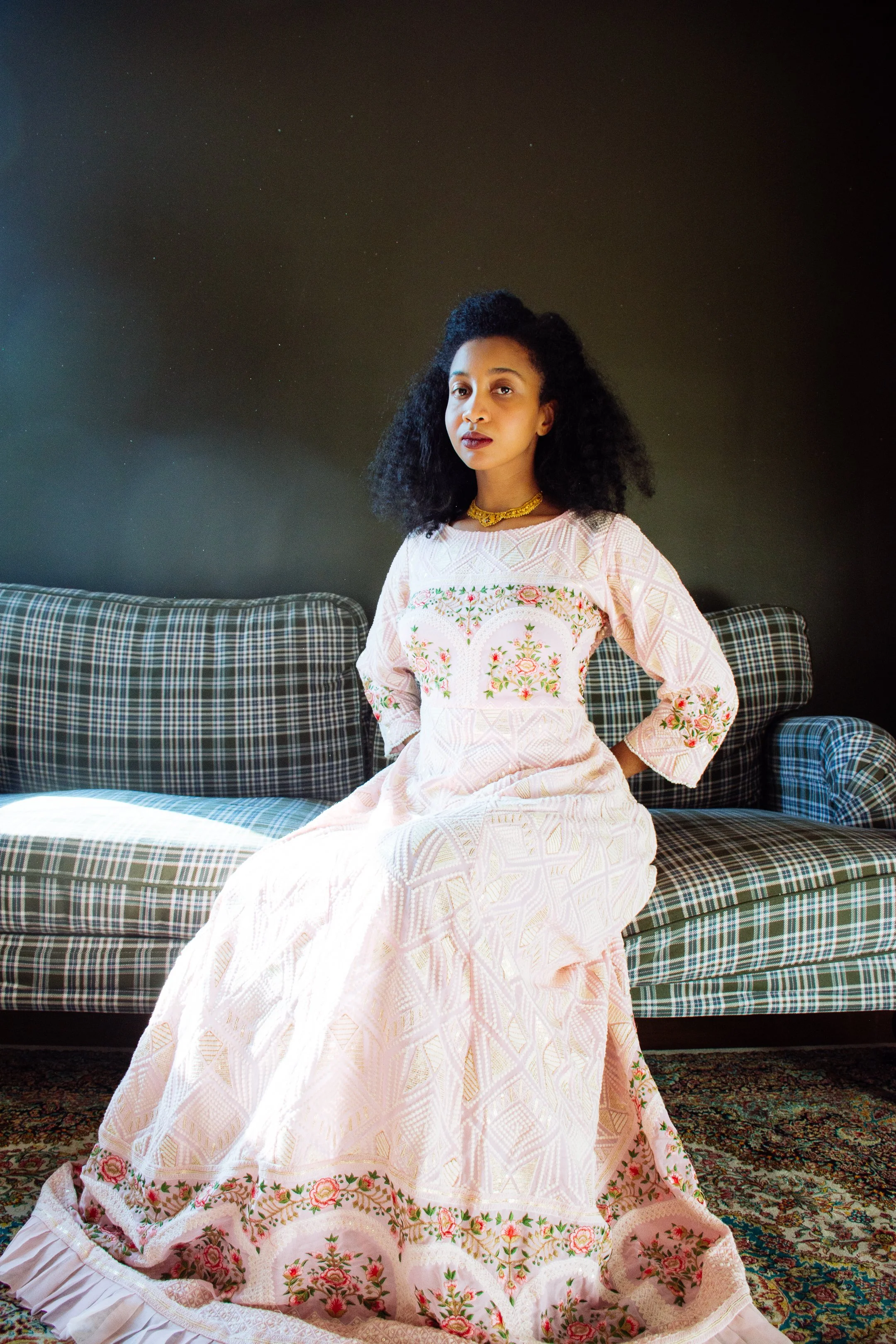 A woman with dark curly hair and dark lipstick sitting on a plaid couch wearing a long pink embroidered dress with floral patterns and gold accessories, in a room with a dark wall, colorful carpet, and plaid furniture.