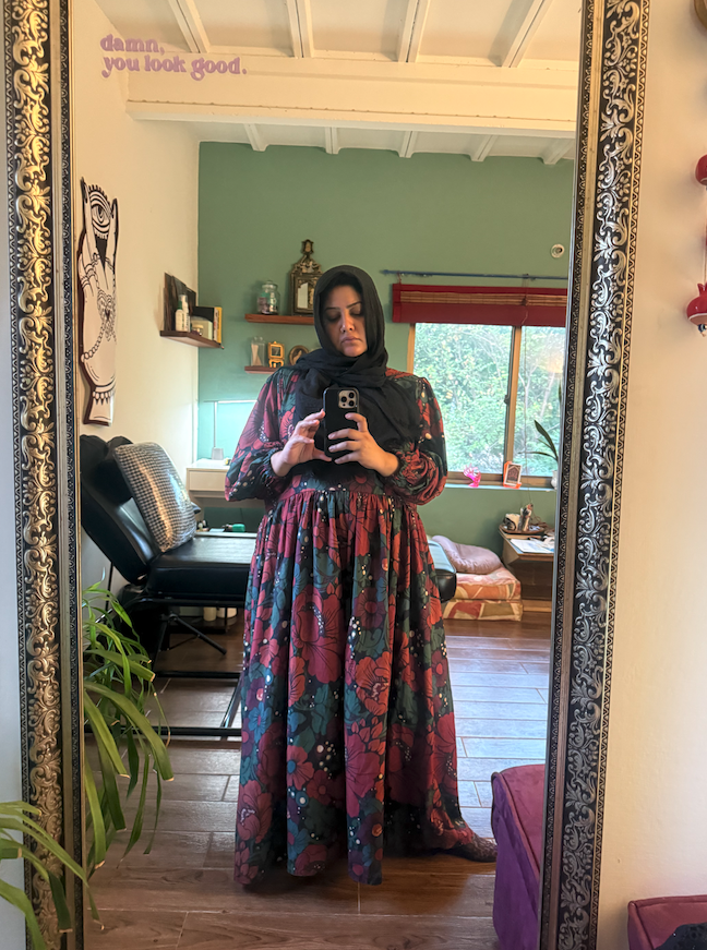 A woman taking a mirror selfie in a room with teal walls. She is wearing a long floral dress with a black headscarf. The room has various decorations, including framed pictures, a desk with a chair, a window with red blinds, and some plants.