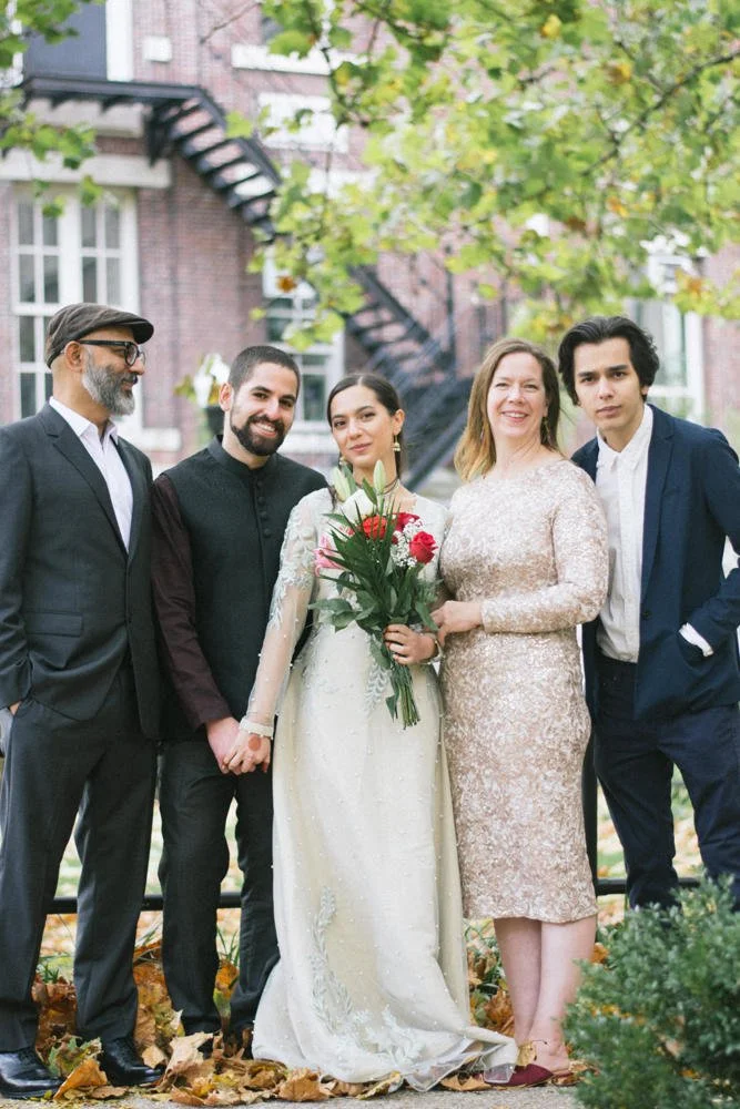 Group of five people standing outdoors on fallen leaves, posing for a photo with a brick building and green tree in the background. A bride in a white dress holding a bouquet of red and white flowers stands in the center, flanked by  men and women in formal attire.