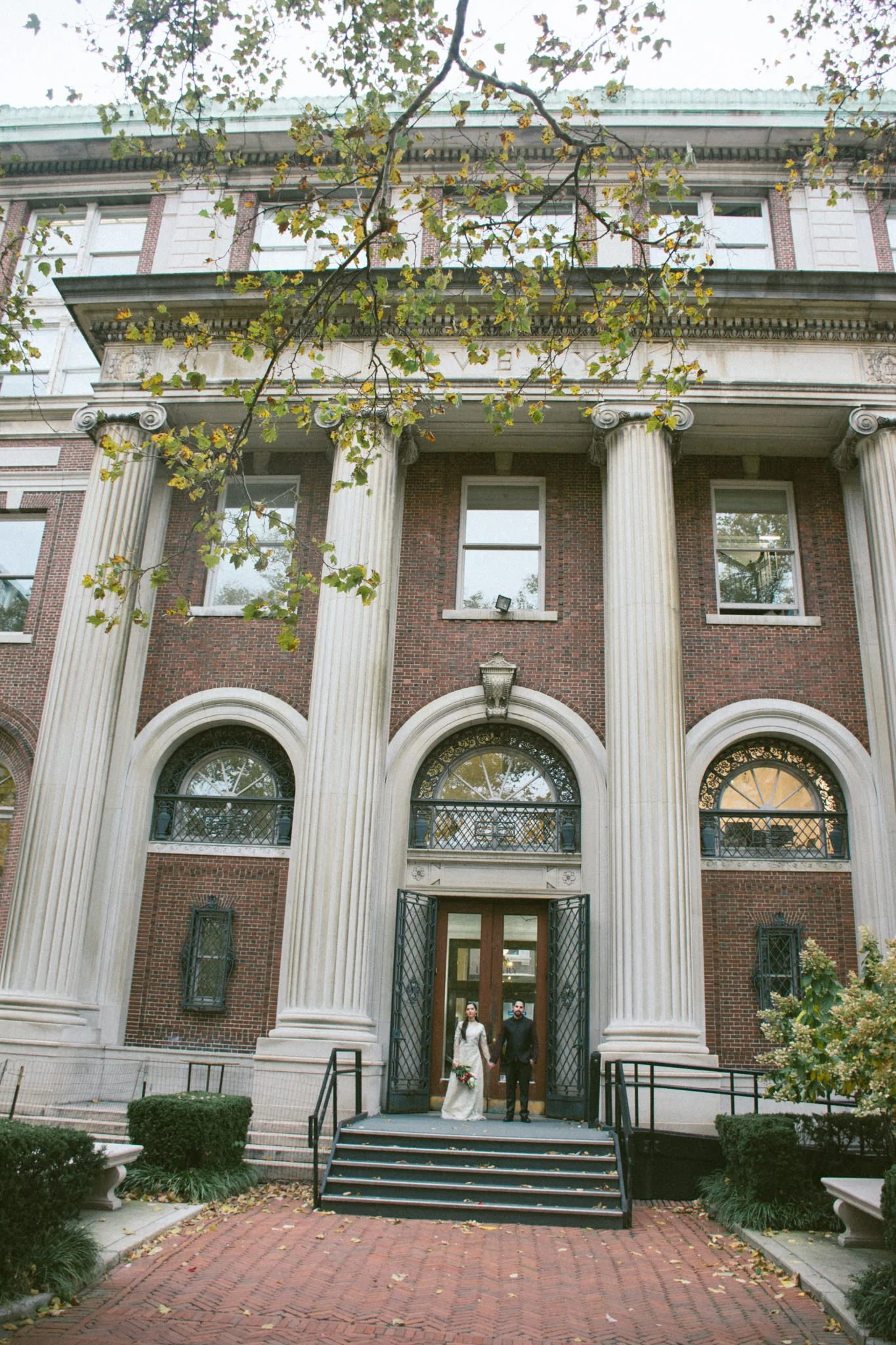 A bride and groom walking out of a historic brick building with large columns, engaged in a wedding ceremony or celebration, holding hands with the bride holding a bouquet of flowers.
