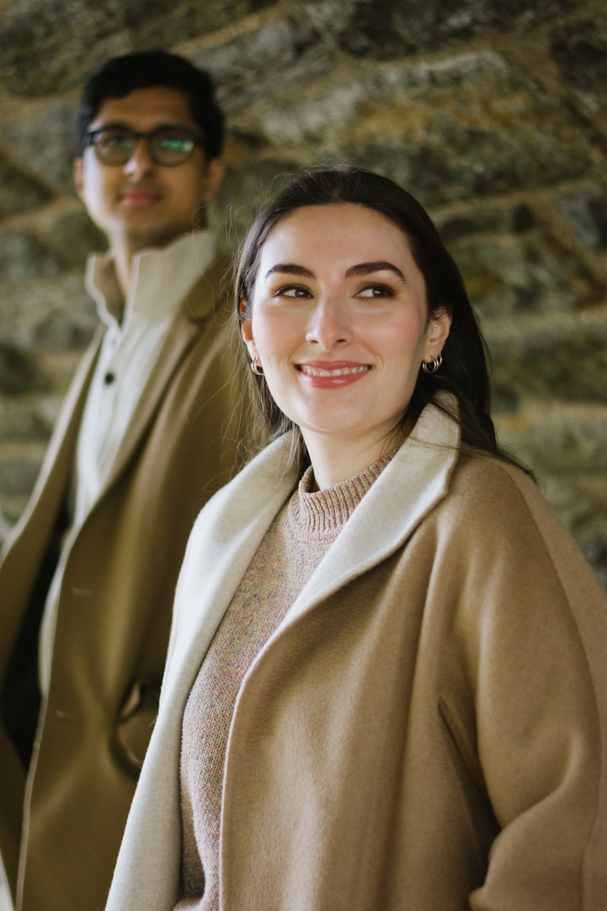 A young woman with dark hair, wearing a beige coat and hoop earrings, smiling and looking to the side, standing in front of a man with dark hair and glasses, also wearing a beige coat, against a rocky background.