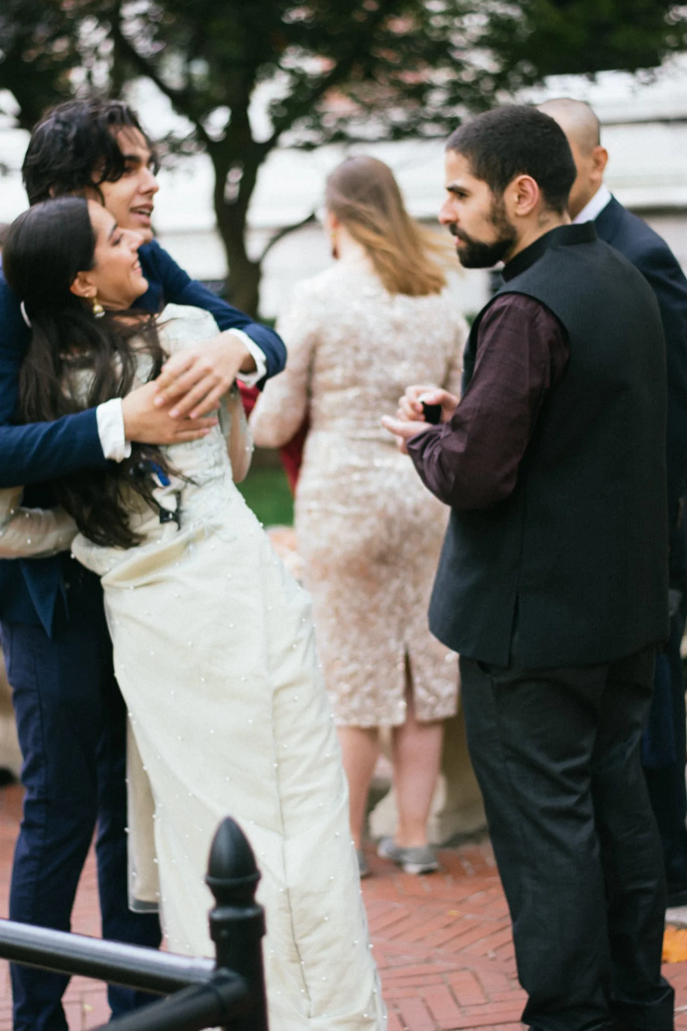 A wedding scene with multiple people, including a woman in a white wedding dress, being embraced by a man in a navy suit. Other guests are visible in the background, with some talking and standing outside on a brick-paved area near a tree.