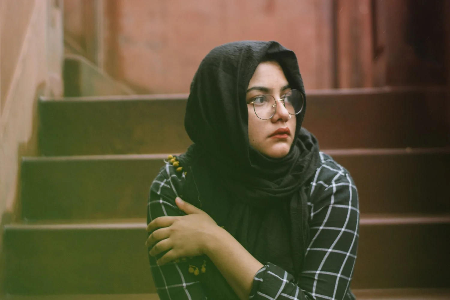 A young woman with glasses, wearing a black headscarf and a black and white checkered shirt, sitting on a staircase, looking to her left with a pensive expression.