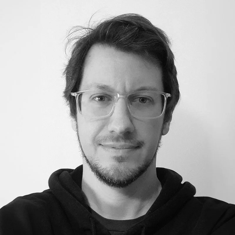 A young man with glasses and short dark hair, smiling slightly, wearing a black hoodie, in front of a plain wall.
