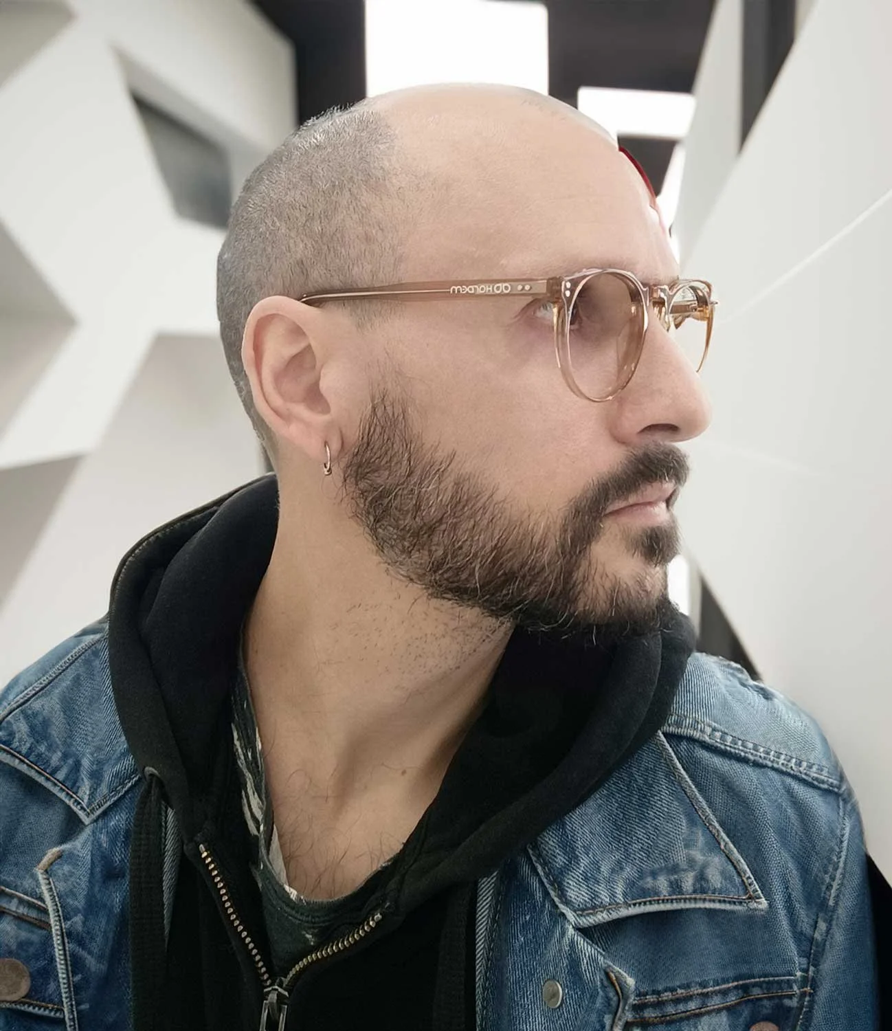 A man with a shaved head and a beard wearing glasses, a hoop earring, a denim jacket, and a black hoodie, looking to the right, with an abstract geometric background.