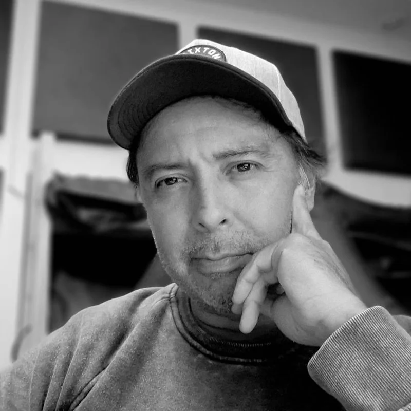 A man with a baseball cap resting his chin on his hand, looking directly at the camera in a black and white photo.