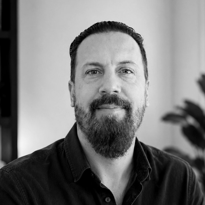 Black and white portrait of a man with a beard and slicked-back hair, wearing a dark collared shirt, looking directly at the camera.