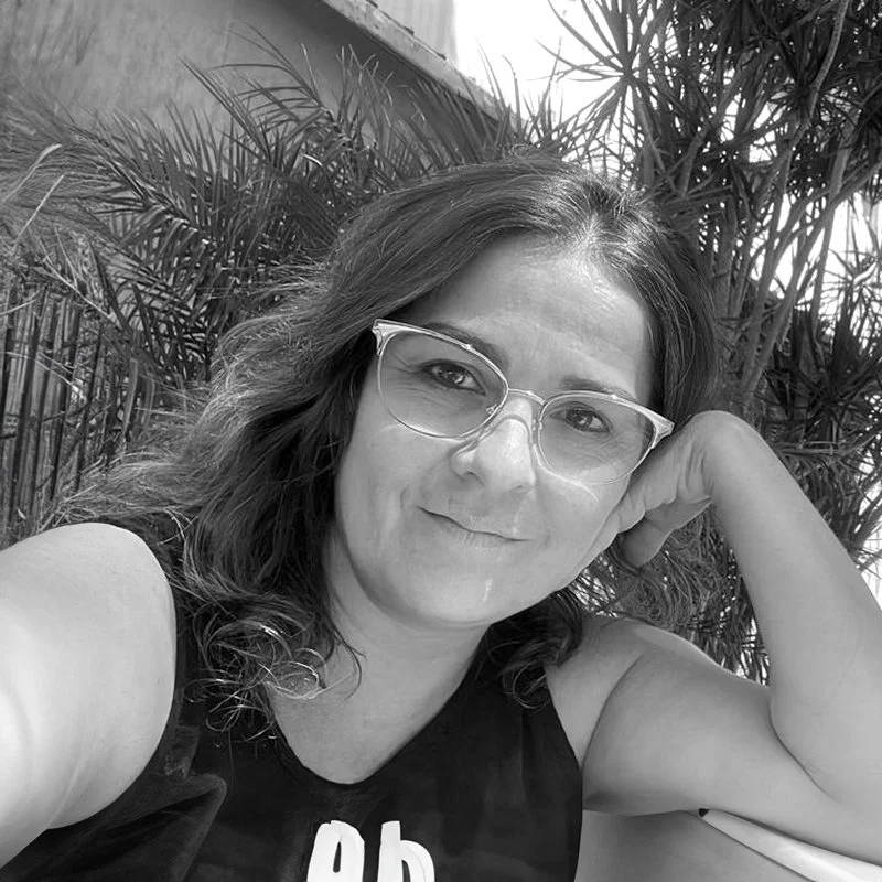 A woman with wavy hair wearing glasses, smiling, resting her head on her hand, seated outdoors near plants.