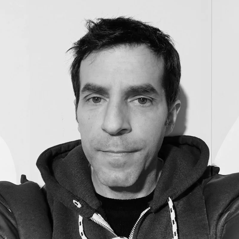 Black-and-white photo of a man with short dark hair, wearing a hoodie with a zipper, looking directly at the camera.
