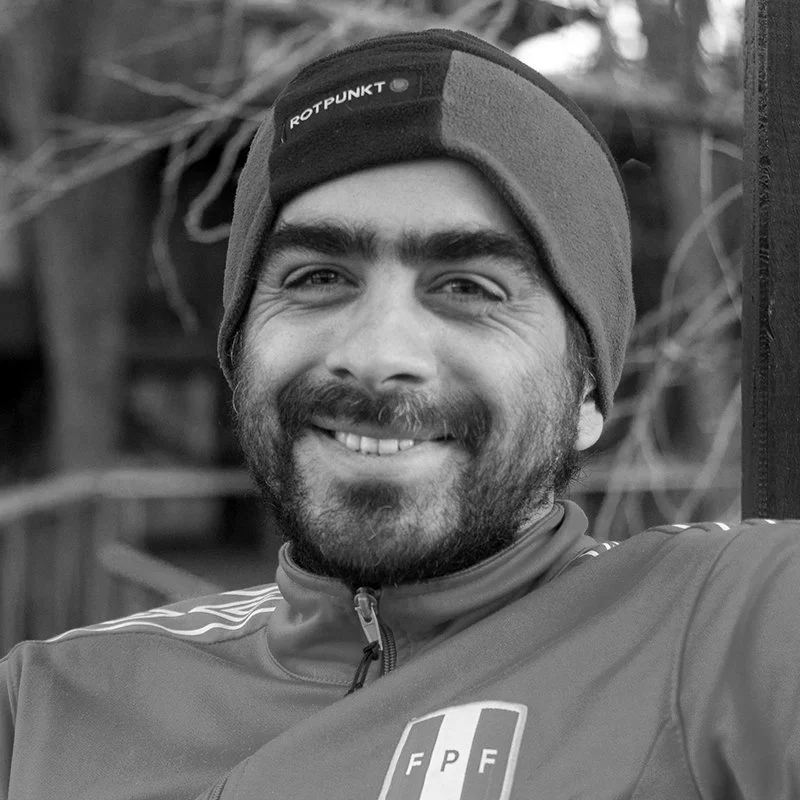 A smiling man with a beard wearing a beanie and a zipped jacket, outdoors with trees and a fence in the background.