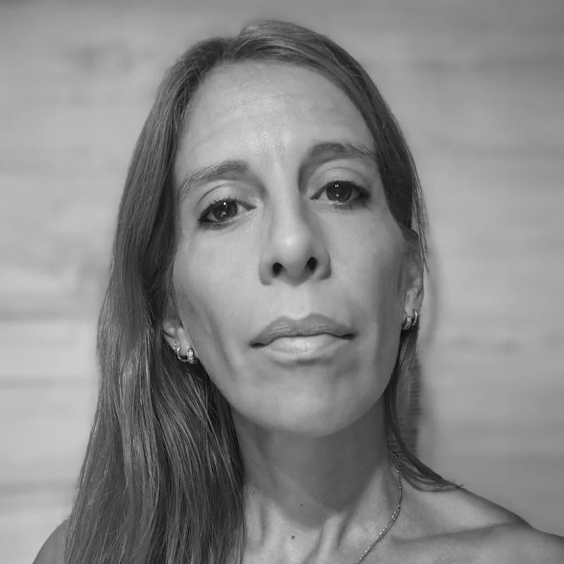 A black and white portrait of a middle-aged woman with long hair, wearing earrings and a necklace, against a blurred background.