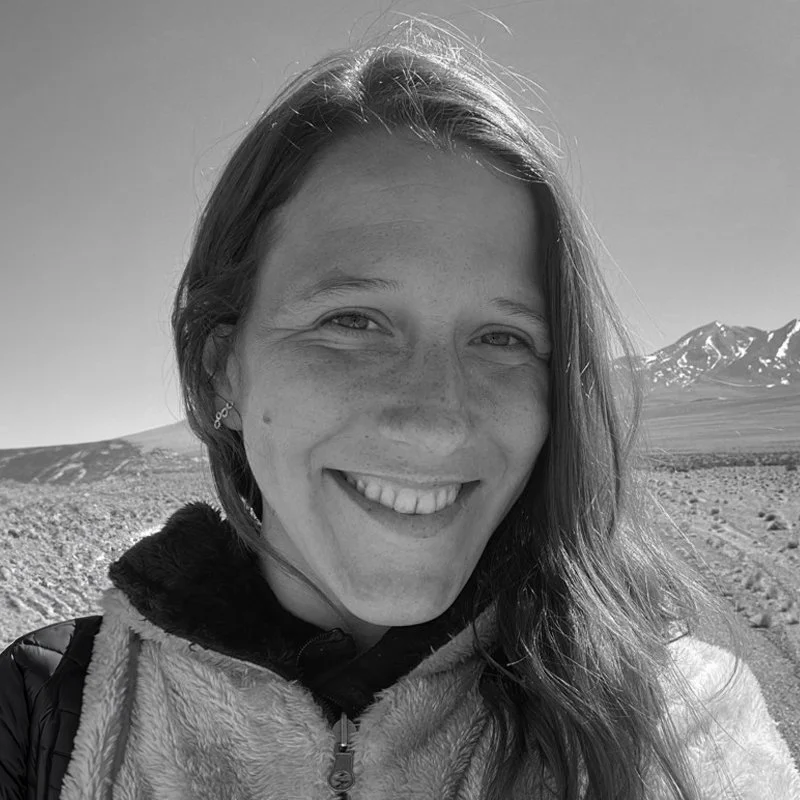A woman smiling outdoors in a mountainous, desert landscape with snow-capped peaks in the background.