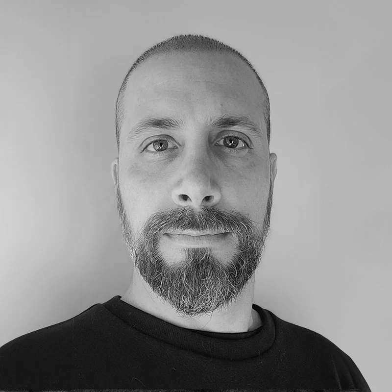 Black and white photo of a man with a beard and mustache, short hair, wearing a dark shirt, looking directly at the camera against a plain light background.