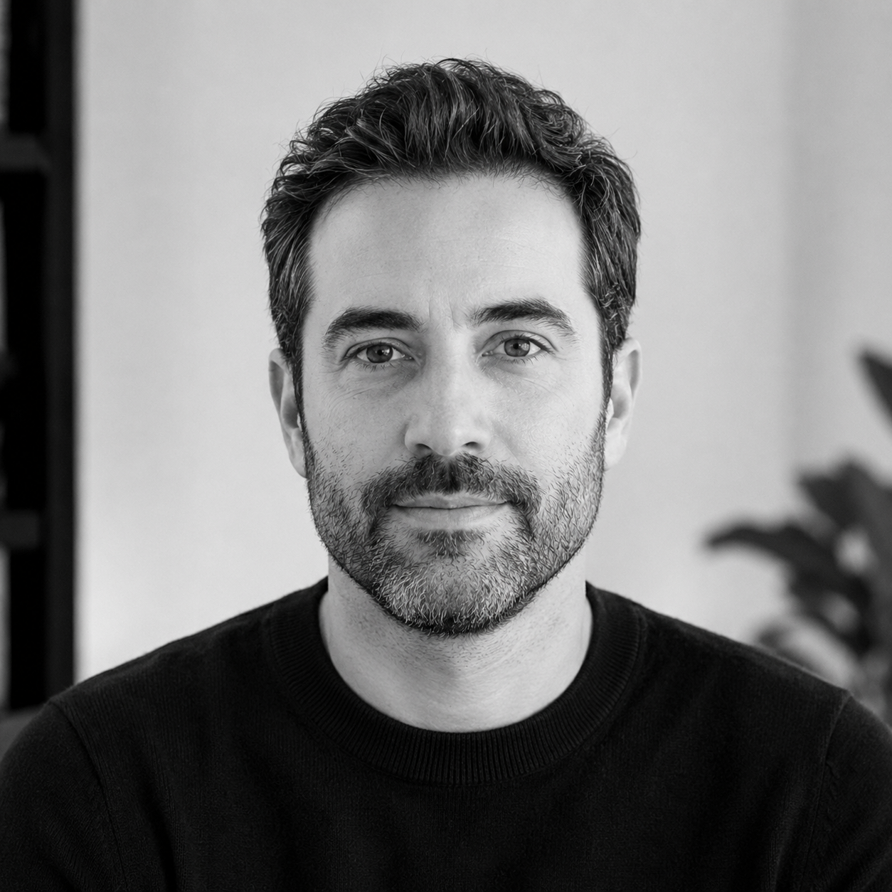 Black-and-white portrait of a man with dark hair and a beard, looking directly at the camera, wearing a black shirt.