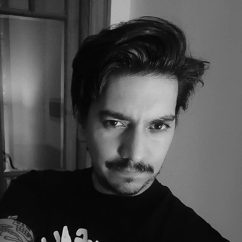 A young man with styled dark hair and a mustache taking a selfie in black and white, looking directly at the camera with a serious expression.
