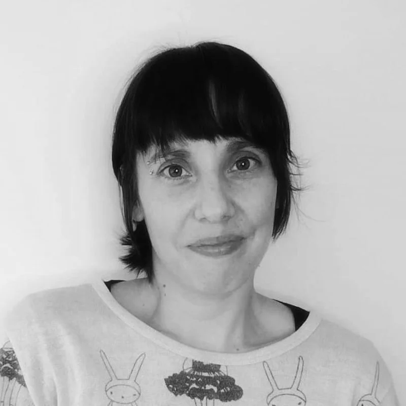 Black and white photo of a woman with short dark hair, wearing a light-colored top with bunny and cloud prints, standing in front of a plain white background.