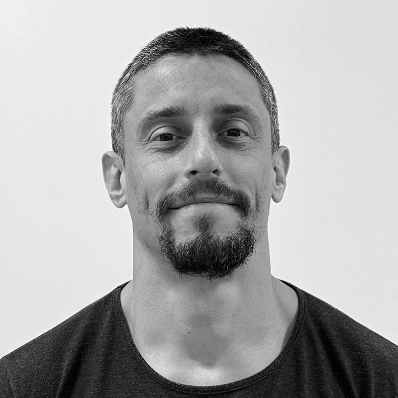 A black and white portrait of a man with short hair, a goatee, and a slight smile, wearing a dark shirt, standing against a plain light background.