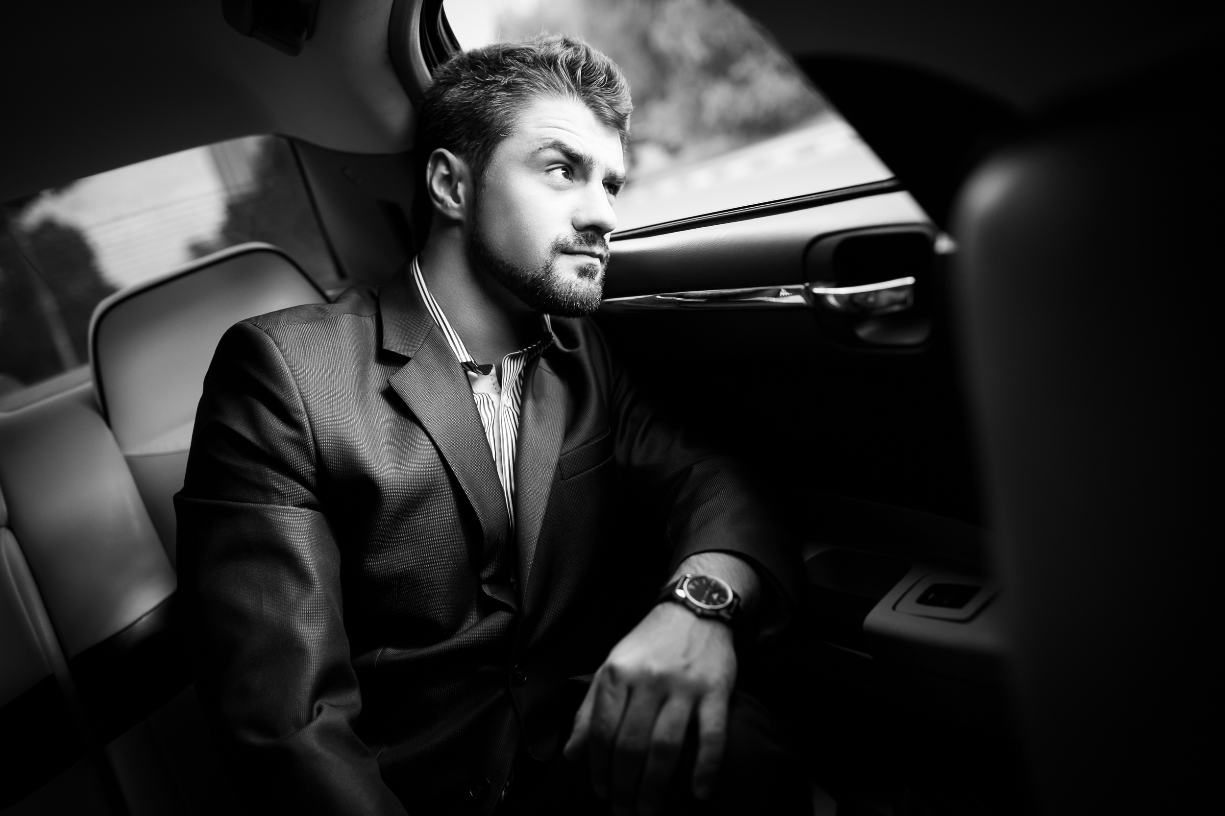 Black-and-white photo of a man in a business suit sitting in the back seat of a car, looking out the window with a thoughtful expression.