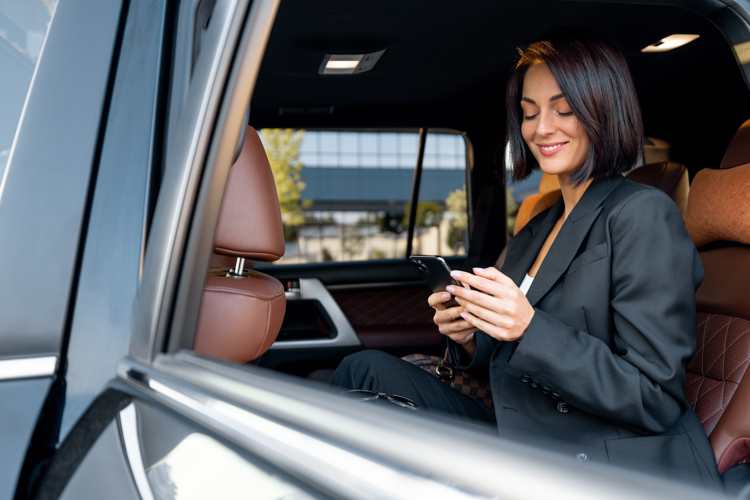 A woman with dark hair sitting in the back seat of a vehicle, using her phone and smiling.