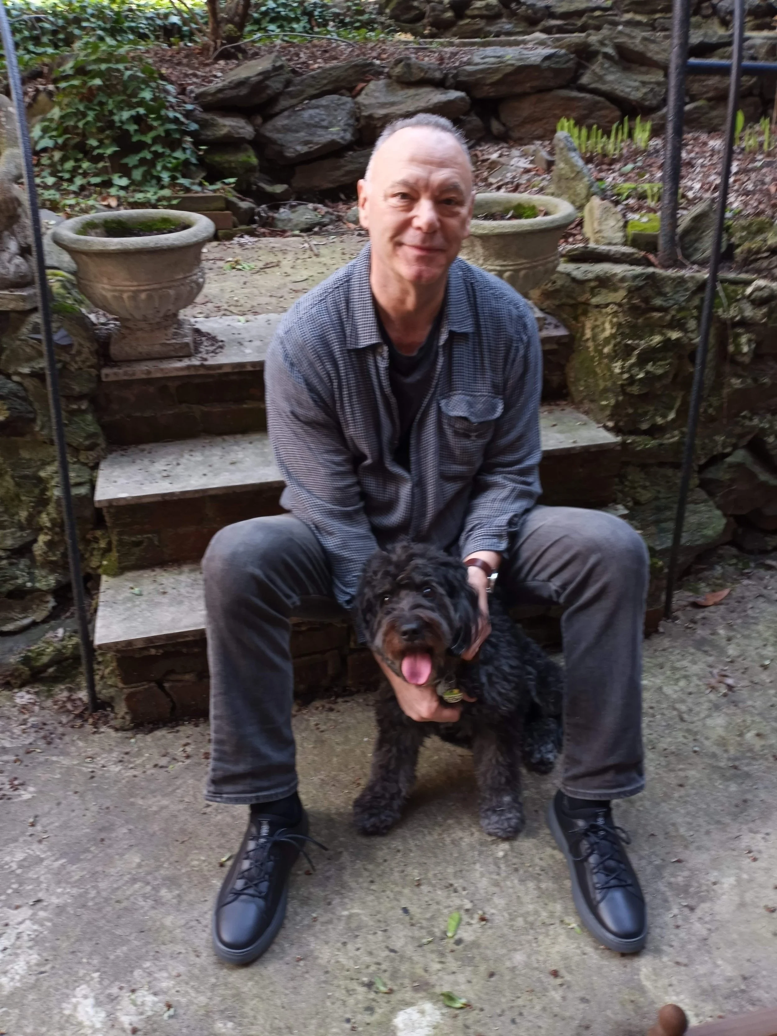 Michael Cannistraci sitting on outdoor steps holding a black curly-haired dog with tongue out, in a garden with stone planters and a stone wall background.
