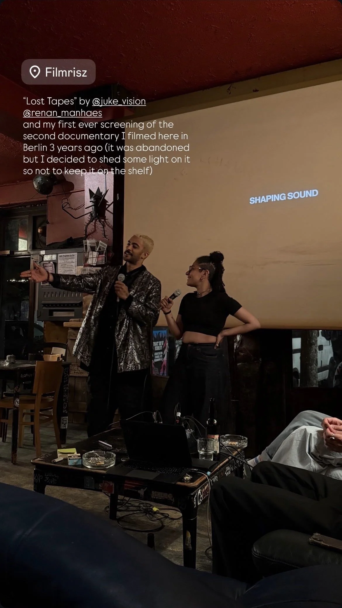 Two performers, one male with blond hair wearing a shiny suit and one female with dark hair in a bun and glasses, stand on stage holding microphones in a dimly lit restaurant or bar. A large screen behind them displays the words "SHAPING SOUND." The audience is seated, with a table in the foreground holding drinks, a laptop, and other items.