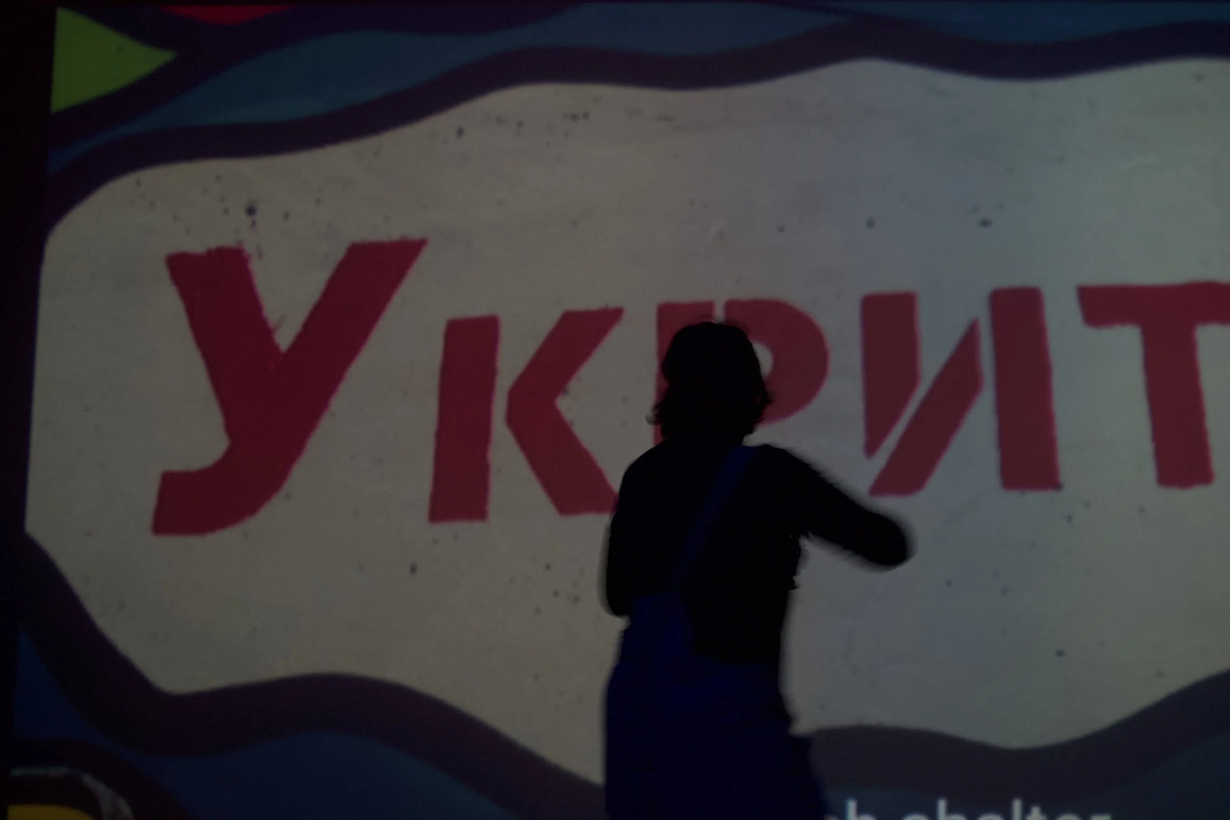 A person with dark hair silhouetted against a brightly lit sign with red text. The sign has a wavy top border with blue, green, and red colors and a white background. The person appears to be standing in front of the sign, partially blocking the view of some of the red text.
