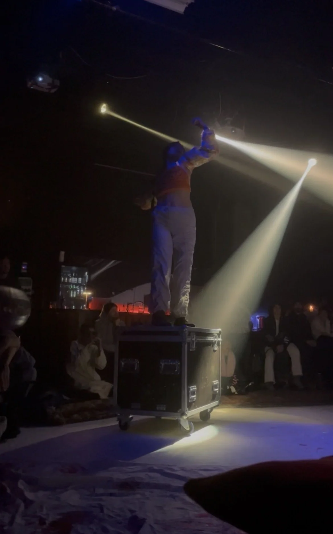 A performer standing on a large equipment case on a stage, holding a flashlight, with a curved line of light around them, in a dark room with an audience watching.