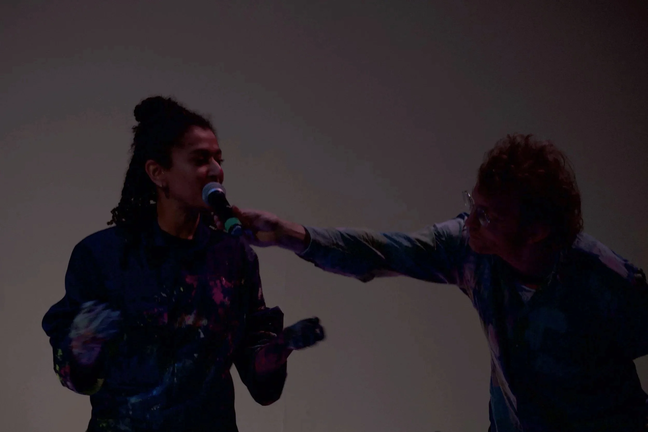 Two people, one with long hair and the other with curly hair, stand on stage under dim lighting. The person with long hair is speaking into a microphone, and the other is reaching out, touching their shoulder.