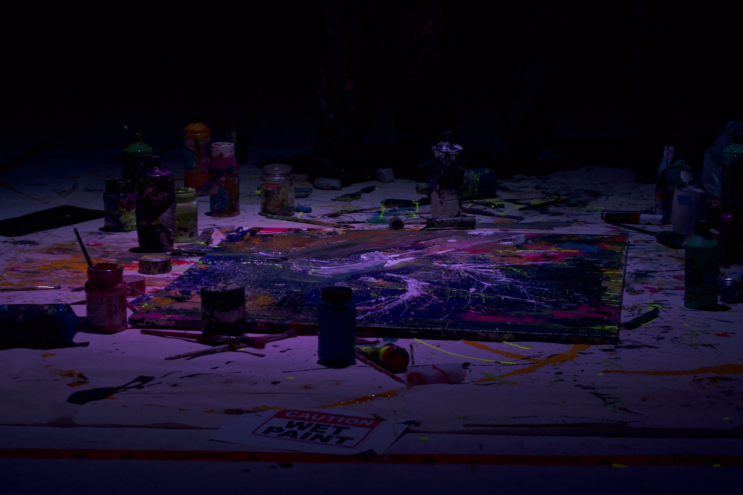 An art studio workspace with paint bottles, brushes, and a paint-splattered surface under blacklight.