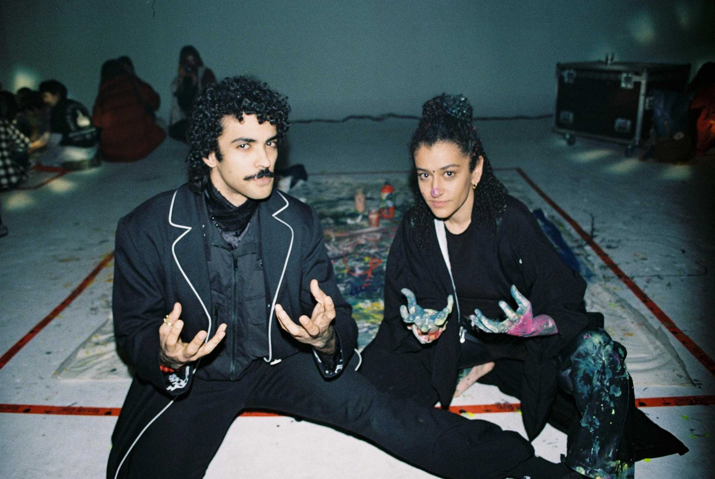 Two people sitting on the floor in a room with other people in the background. They are posing for the camera, with one person making a gesture with their hands and the other with painted hands and a painted face.