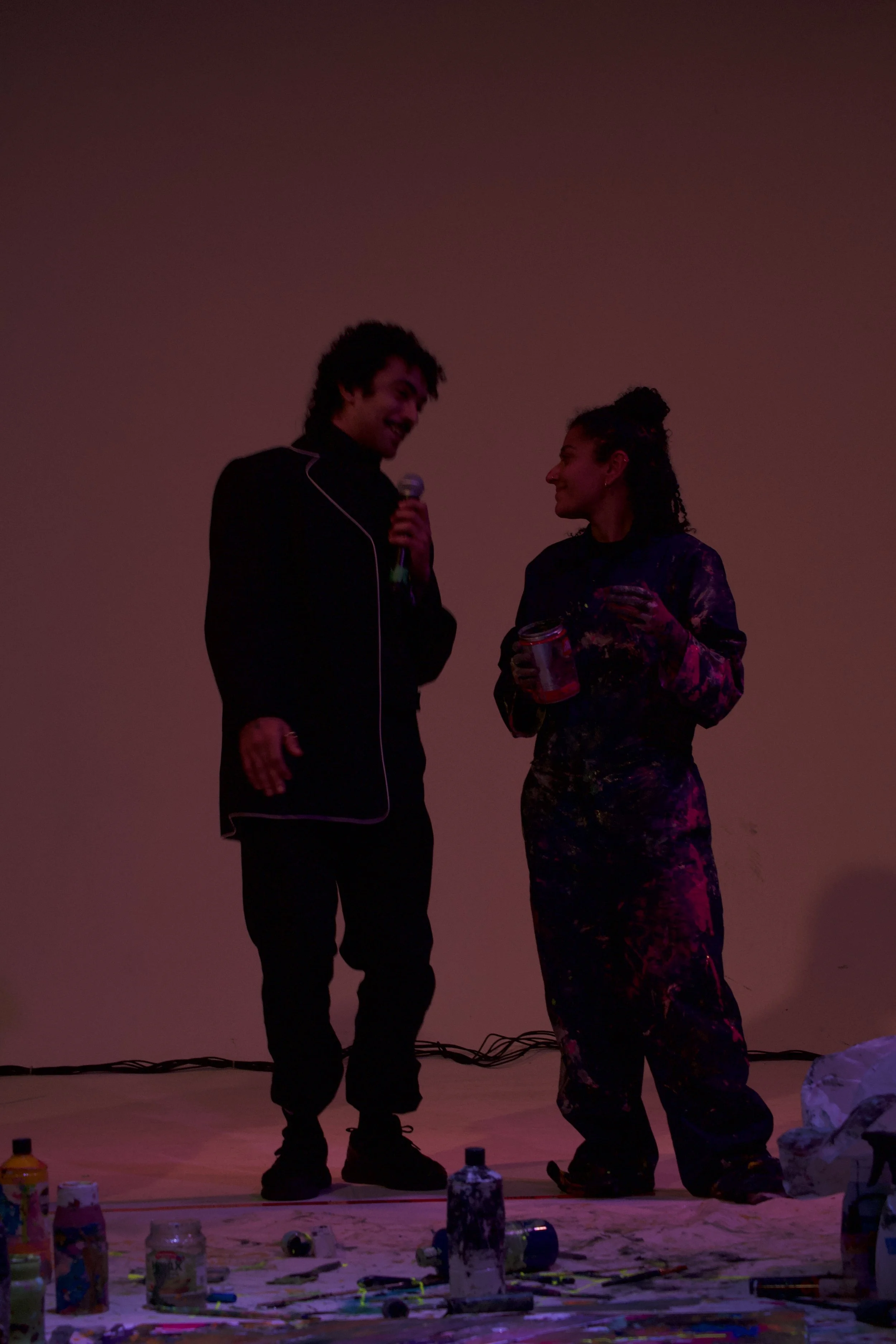 Two people stand on a stage, one holding a microphone, with art supplies and spray paint cans on the floor. They are smiling at each other as they converse.