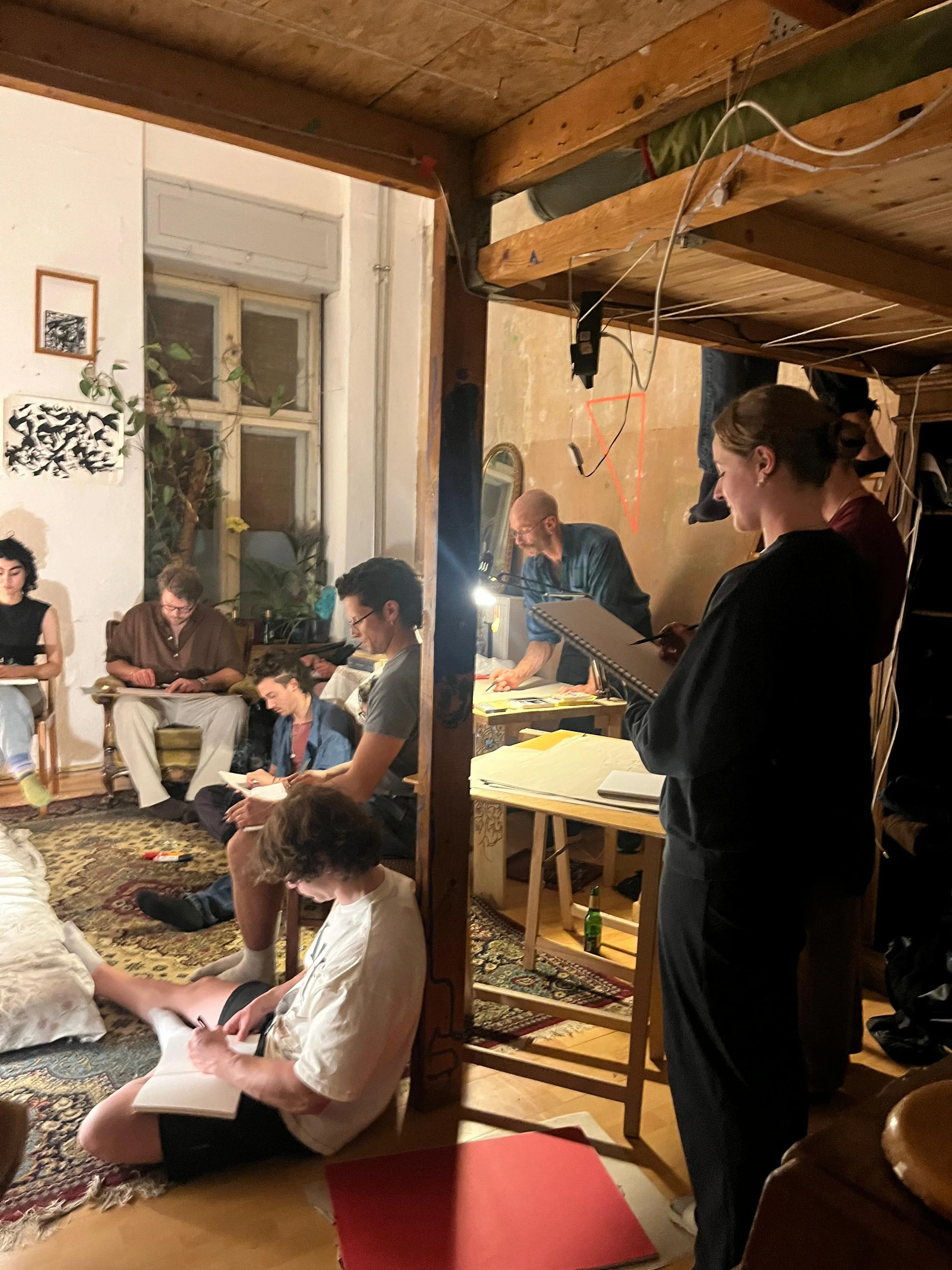 People gathered in a cozy room with warm lighting, sitting and standing around, some engaging with books or notebooks, in an informal gathering or meeting.