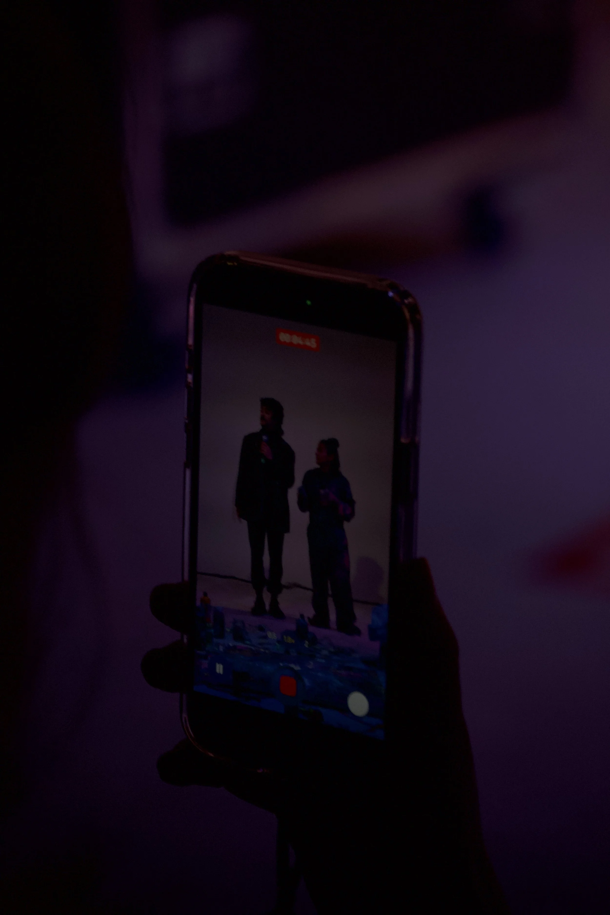 A person holding a smartphone captures a photo of two people standing on a stage, with a dark background and purple lighting.