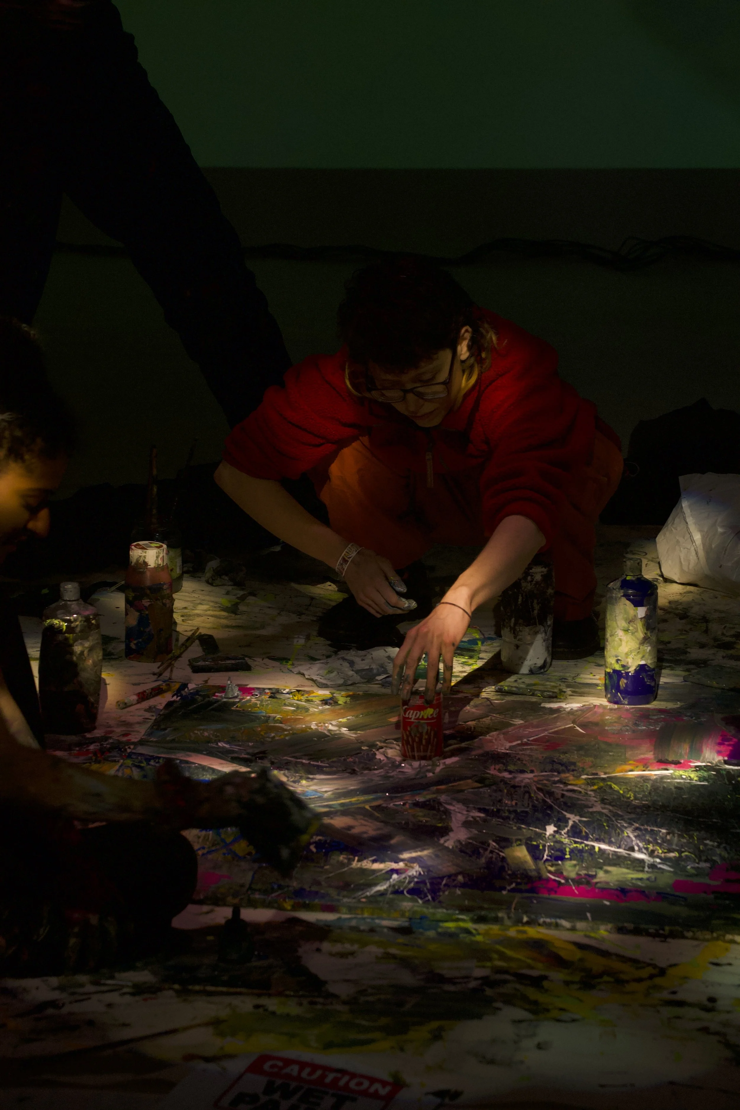 Person in a red hoodie doing a paint splatter art project on a large canvas on the floor, with jars of paint and brushes around.