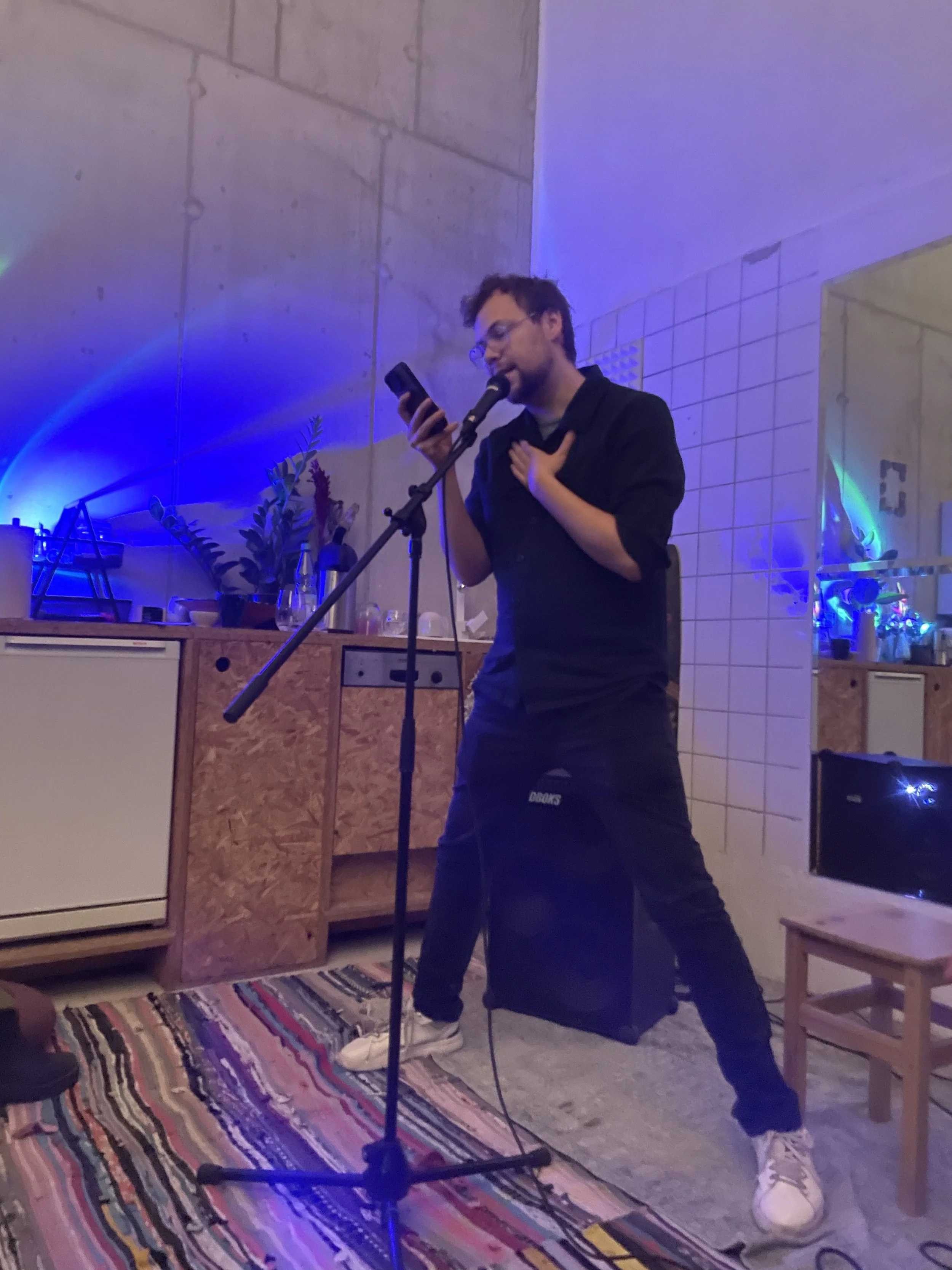 A man wearing glasses, a black shirt, black pants, and white sneakers is performing with a microphone on a stand, holding a phone in his right hand, and placing his left hand on his chest. The room has a concrete wall, a colorful striped rug, and two wooden chairs; there are plants and decorative lights in the background.