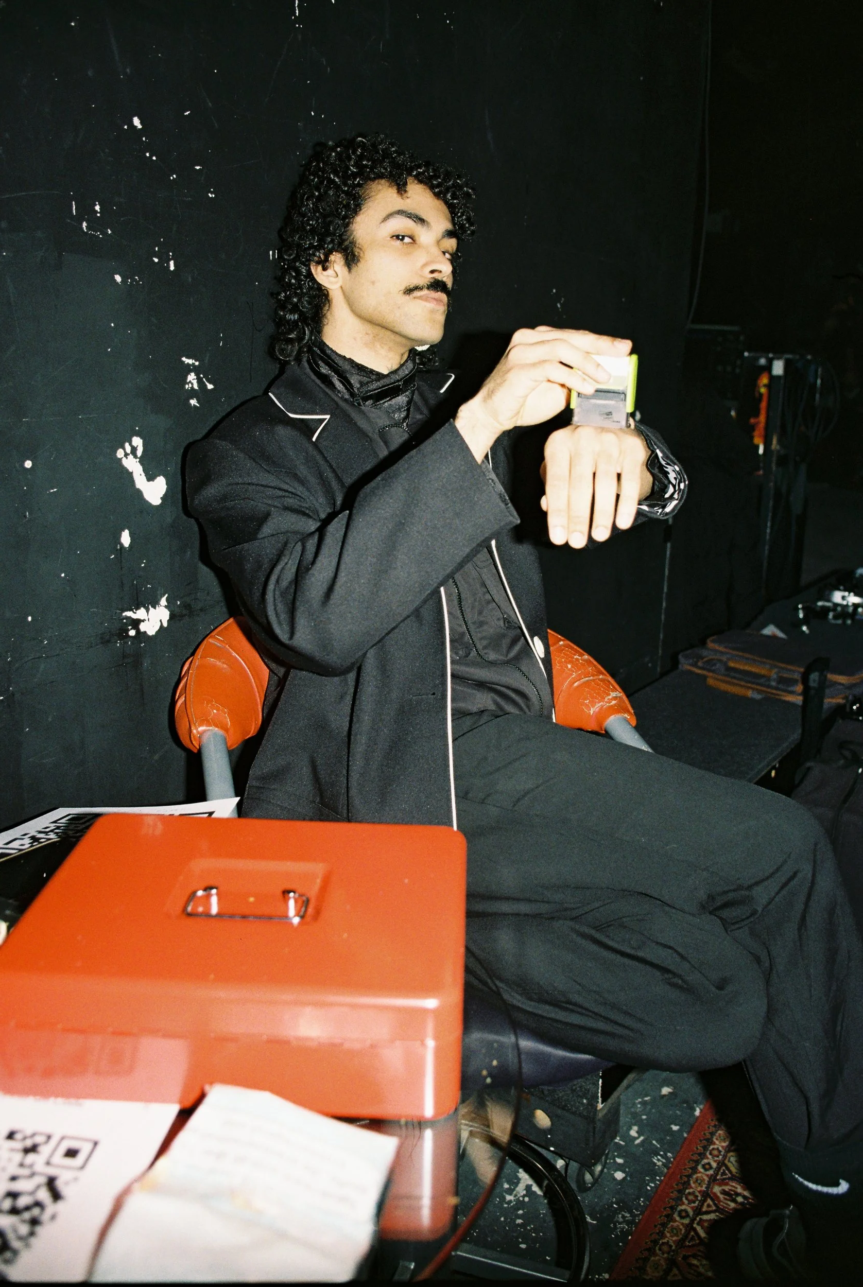 A man with dark curly hair, a mustache, and dressed in a black suit is sitting on a chair. He is looking off to the side and holding a small object, possibly a camera or device, in his hands. There is a black wall behind him, a red box with a latch and a QR code in the foreground, and additional objects and a patterned rug on the floor.