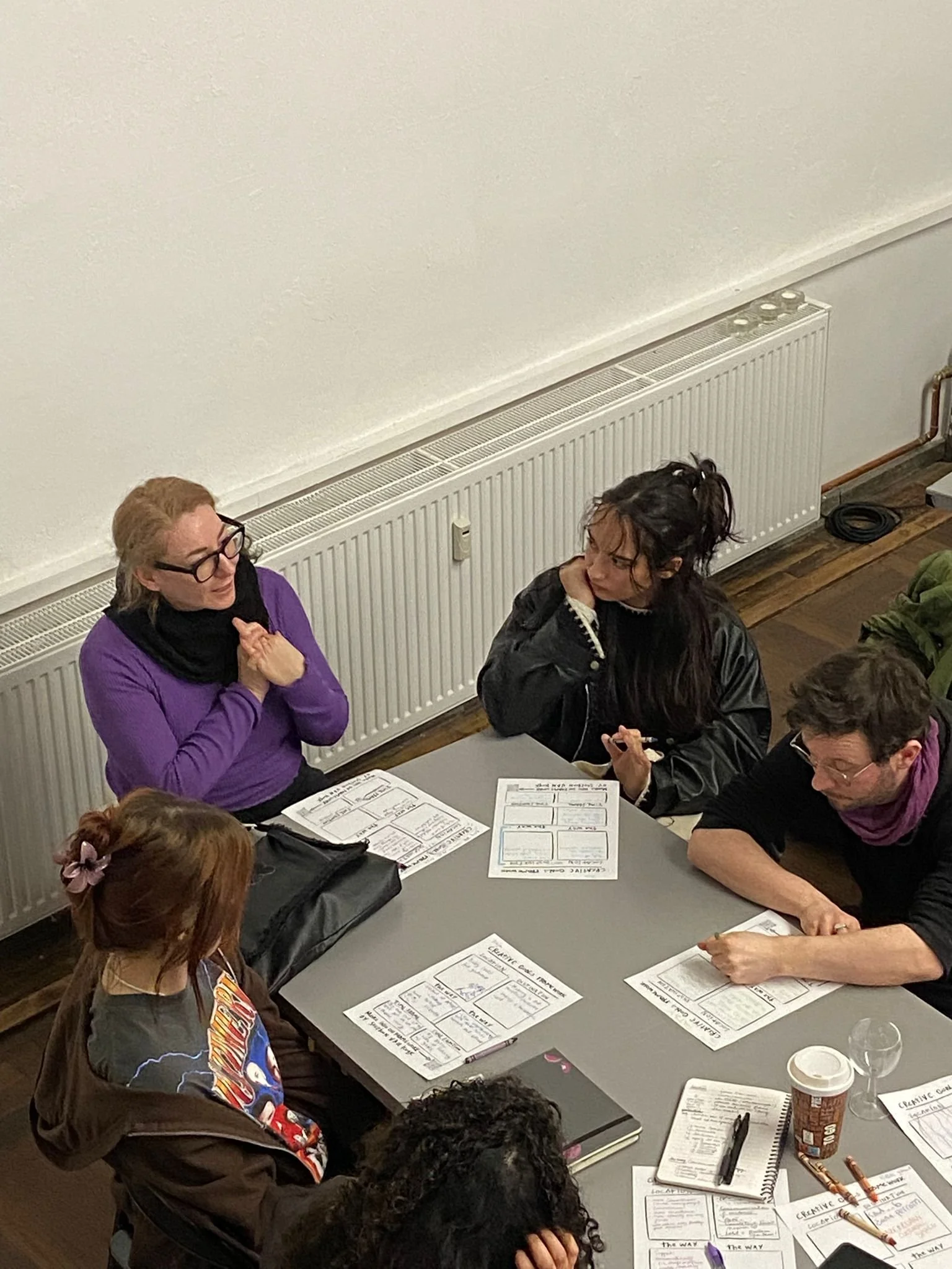 People sitting around a table having a discussion with printed documents, pens, and notebooks in a room with a white wall and a radiator.