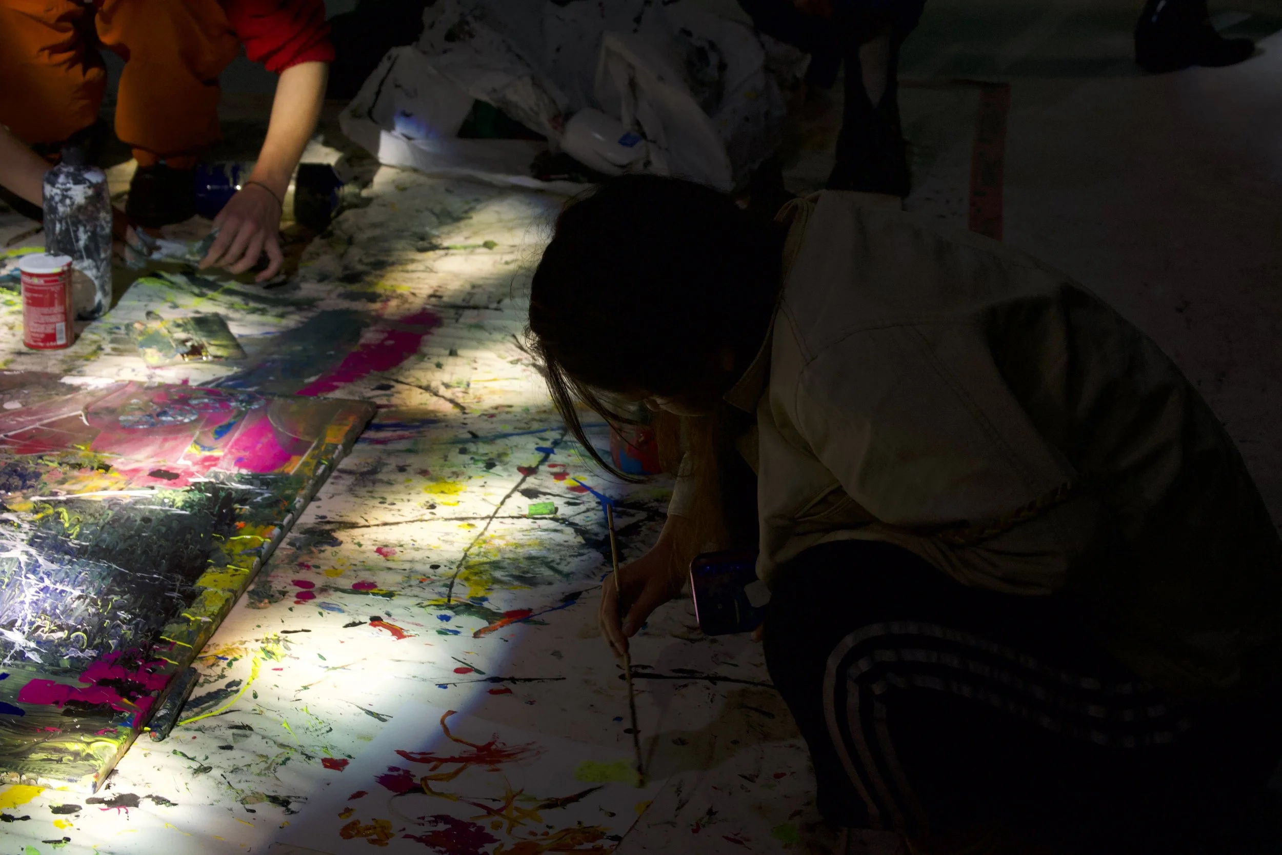 People creating or viewing abstract artwork or painting on a large surface, with colorful paint splatters and strokes, in a darkened room illuminated by focused light.