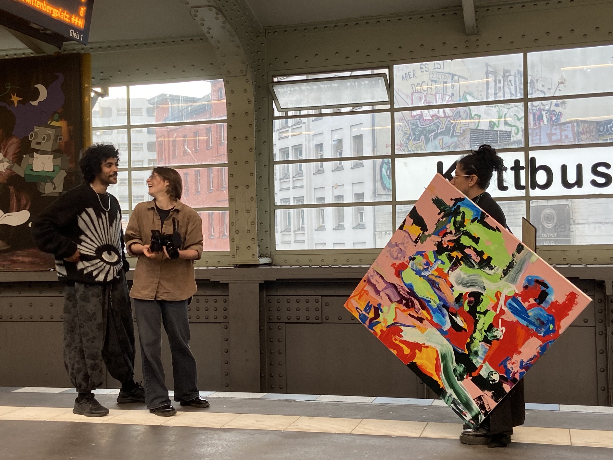 Two people standing and talking in a transit station with large windows and graffiti outside. One person holds a black camera. A colorful abstract painting is in the foreground.