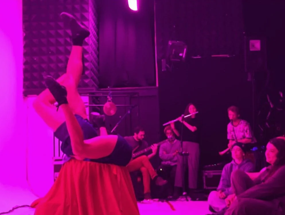 A performer on a table with their legs in the air, with a group of musicians and audience members watching in a dimly lit room with purple lighting.