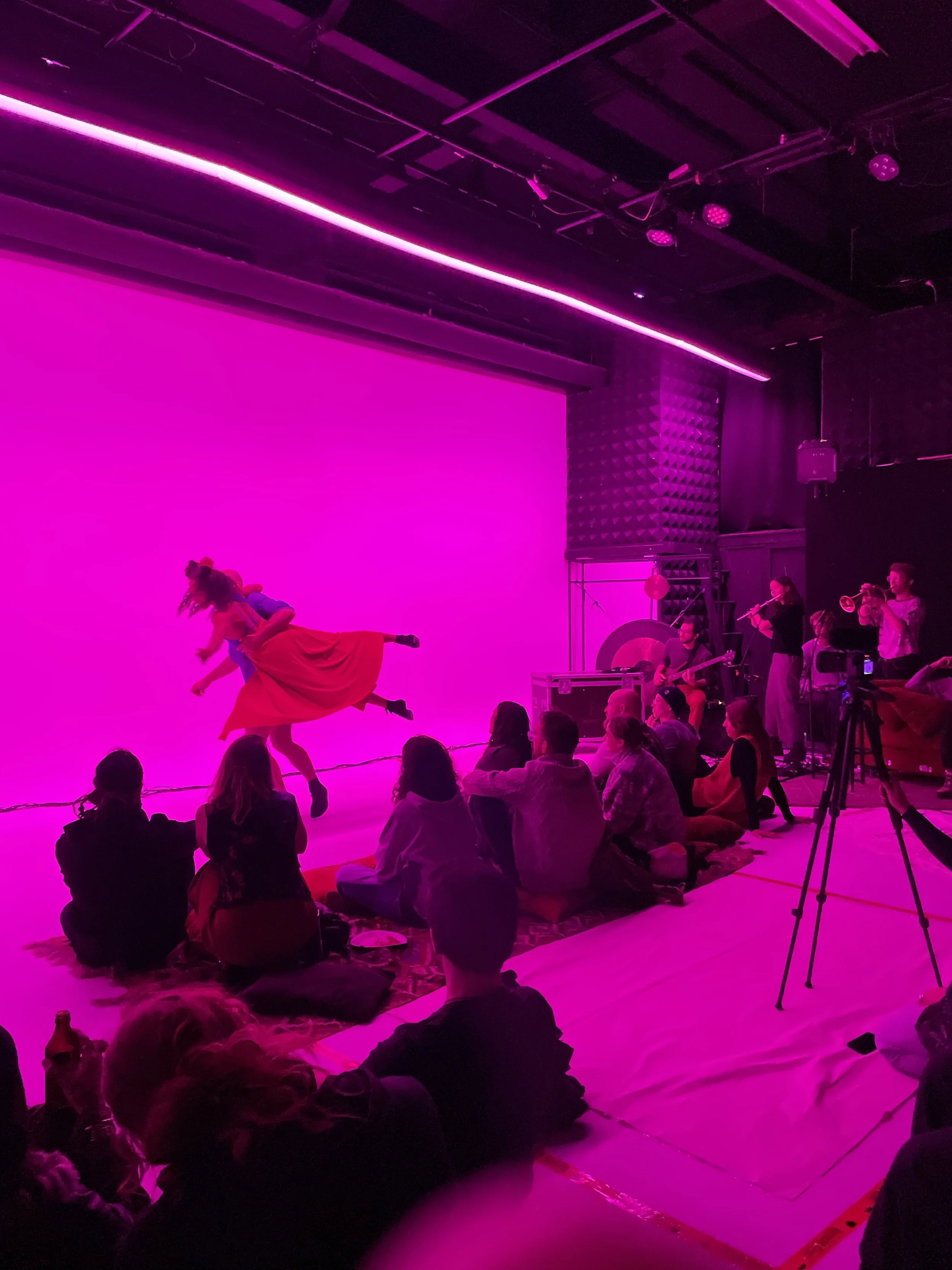 A stage with pink lighting features a group of performers, including a dancer mid-air in a dress. Audience members sit on the floor facing the stage, with some recording the performance. Musical instruments and performers are visible on the right side of the stage.