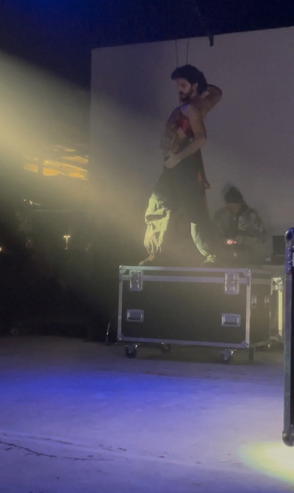 A dancer with dark hair and a beard stands on a raised platform, smiling and looking down. The scene appears to be indoors with stage lighting and a dark backdrop.