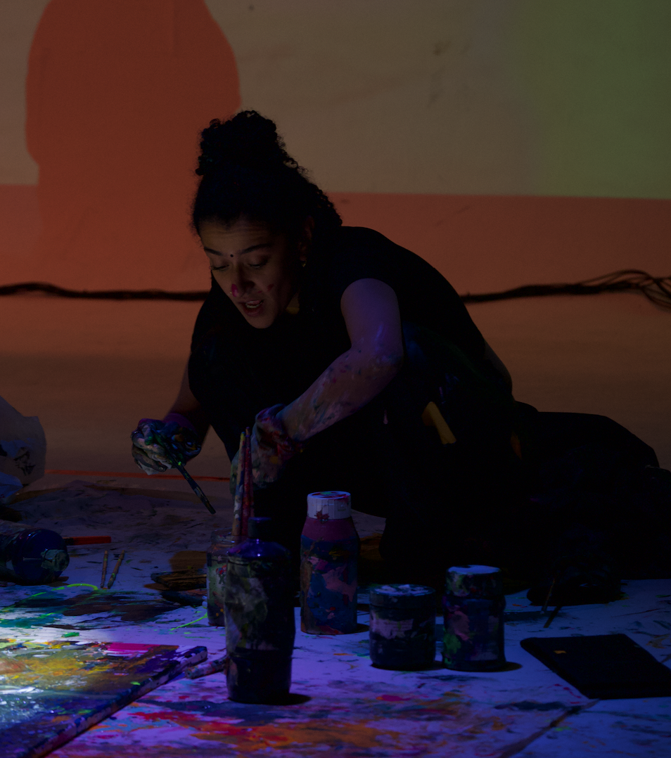 An artist painting with colorful paints on a canvas in a dimly lit room, surrounded by paint bottles and brushes.