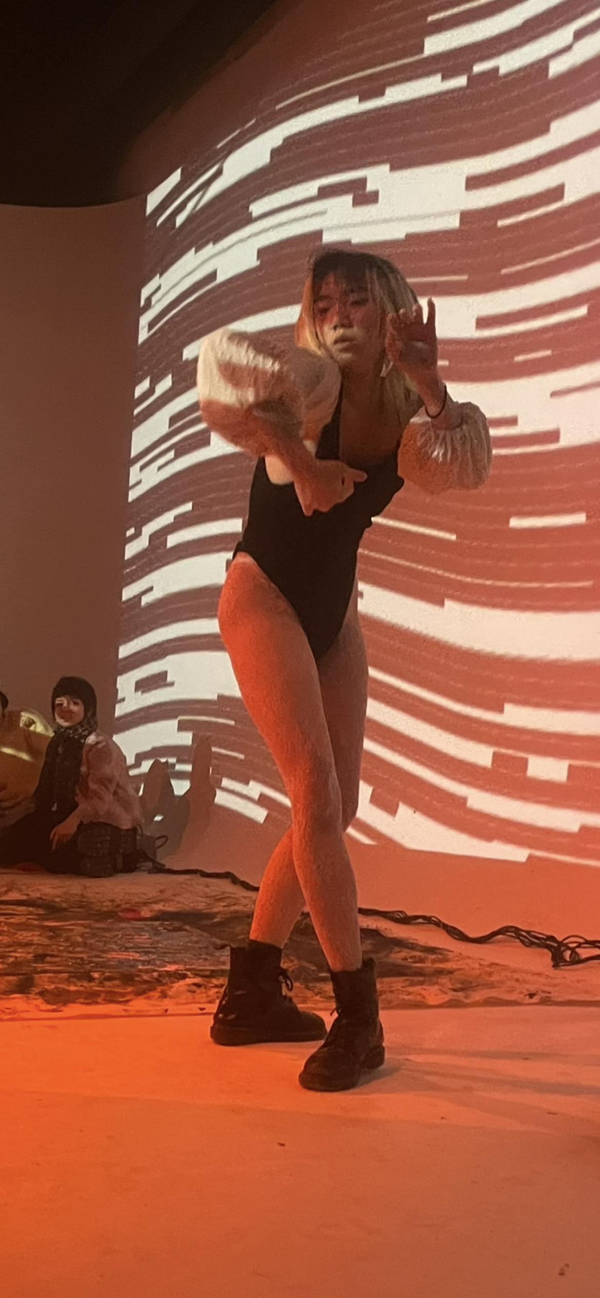 A person standing on stage with a red and white digital pattern projected behind them, wearing a black bodysuit, yellow tights, and black boots, holding a white object.