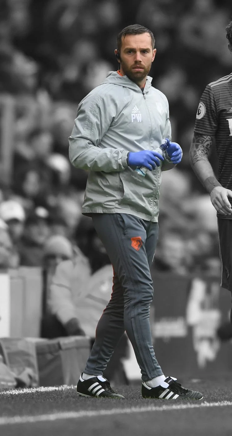Peter Webb, dressed in athletic clothing, stands on the sidelines of a sports field, holding a water bottle and wearing blue gloves. The background is blurred with spectators.