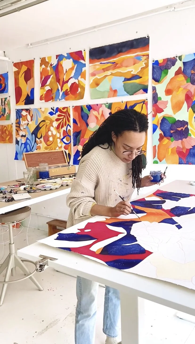 Une artiste peint en utilisant des pinceaux et de la peinture bleue sur une grande toile posée sur une table. Le mur derrière elle est orné de plusieurs œuvres d'art colorées et abstraites.